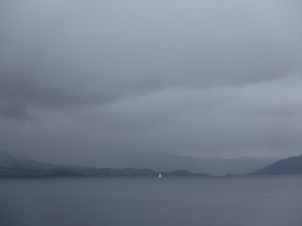 A grey Scottish landscape showing from bottom to top: water on which a white sailboat sails closer to the other shore; hills of the far coast; mist covered mountains stretching out into the distance behind them; and grey clouds hanging over everything.